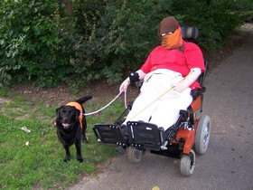 Joyce buiten met haar hulphond Thirza in haar elektrische rolstoel met doeken om haar hoofd voor de pijn